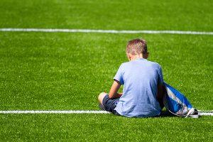 Young white boy sitting on soccer field alone, view from behind.