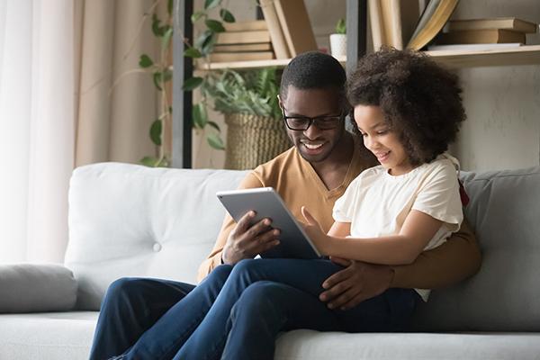 Black father and daughter sitting on a couch smiling at a tablet.
