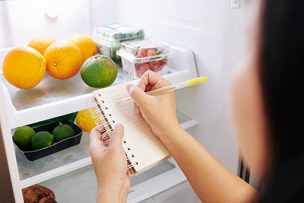 Woman making a list while looking in an open fridge.