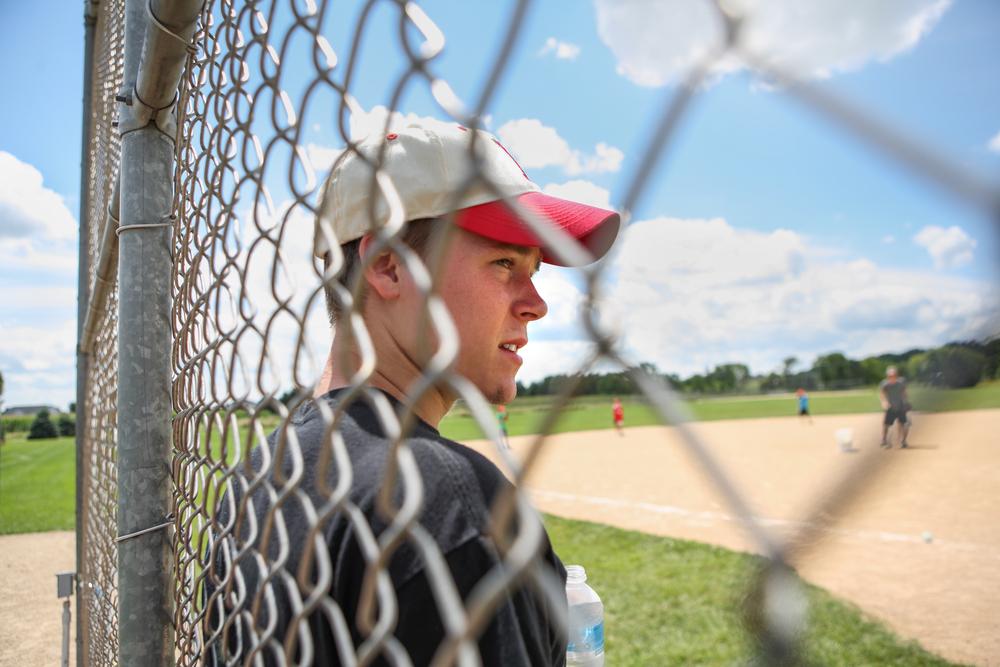 White male teen standing against fence during baseball game.