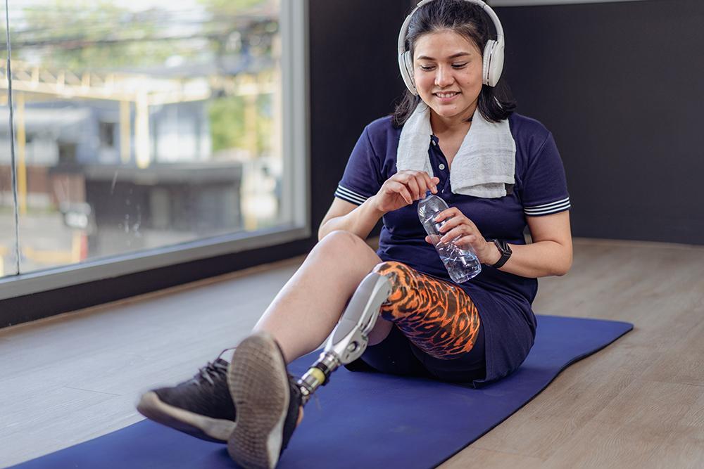Young woman with prostethic leg on yoga mat in gym wearing headphones.
