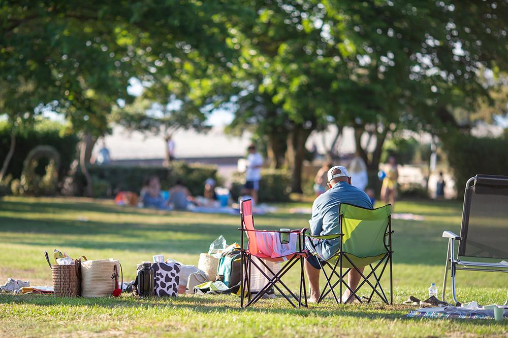 Parent sitting on sidelines outside with coolers and bags.