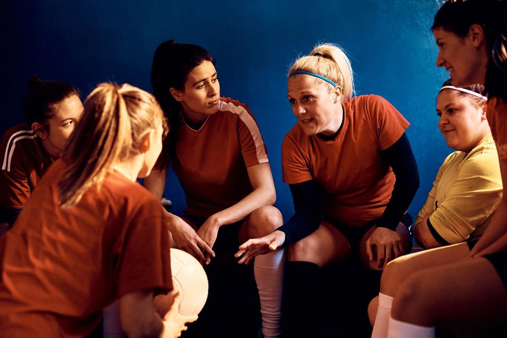 Female soccer players talking amongst themselves on a bench indoors.