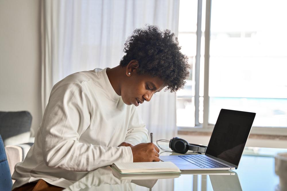 Young male teen writing in a notebook at a desk.