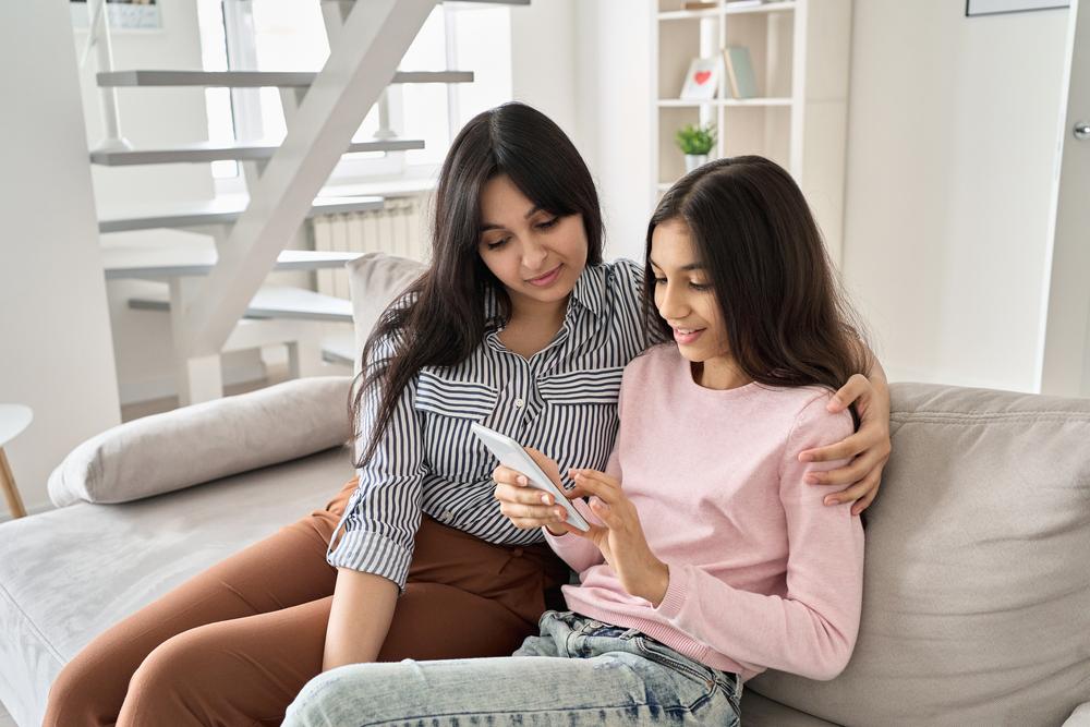 Mother and teen daughter talking on a couch while daughter looks at phone.