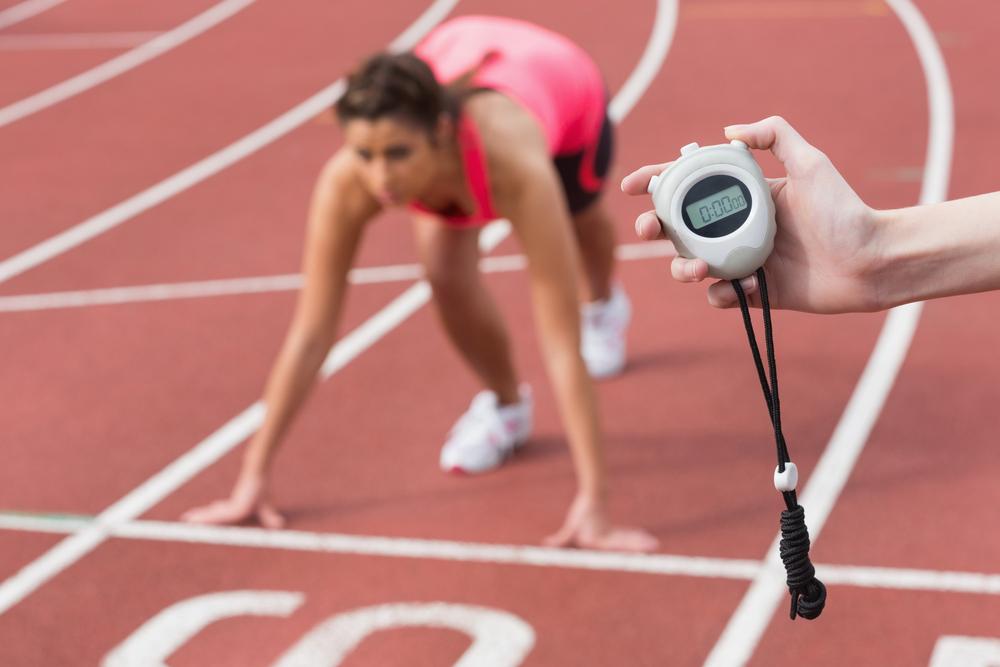 Hand holding a stop watch with a young woman on a track behind the hand.