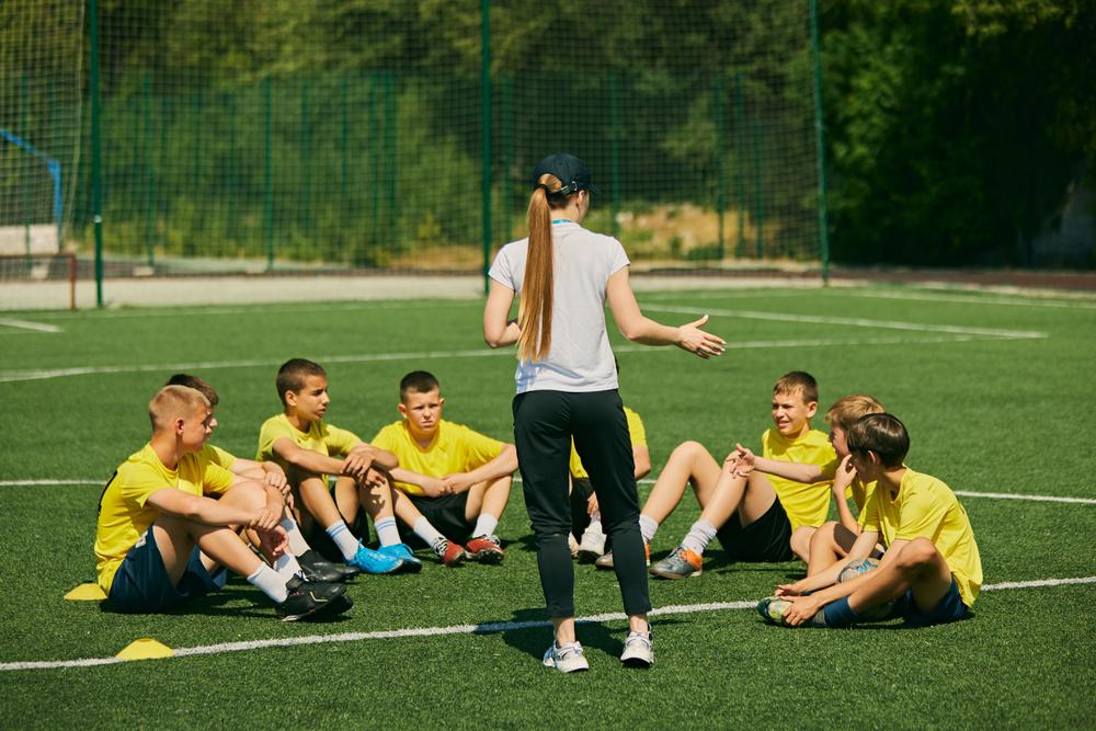 Female coach talking to young male soccer team on field.