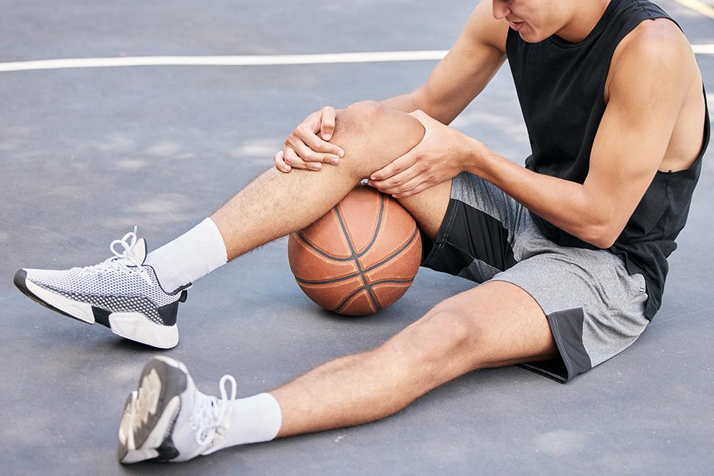 Young man holding area around knee on basketball court with ball.
