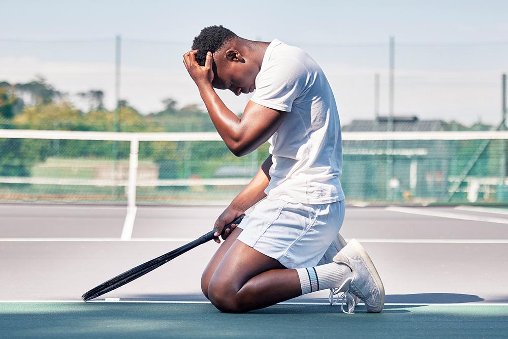 young male tennis player kneeling with racket with head in hands on court.