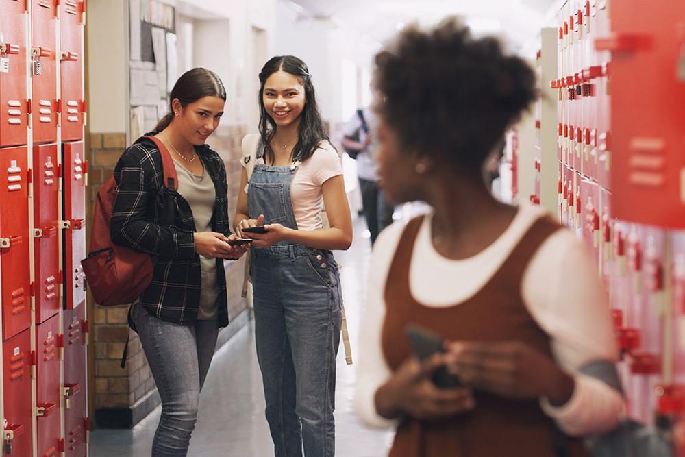 Two teen girls in a high school hallway laughing with each other while looking at a third girl standing further away.
