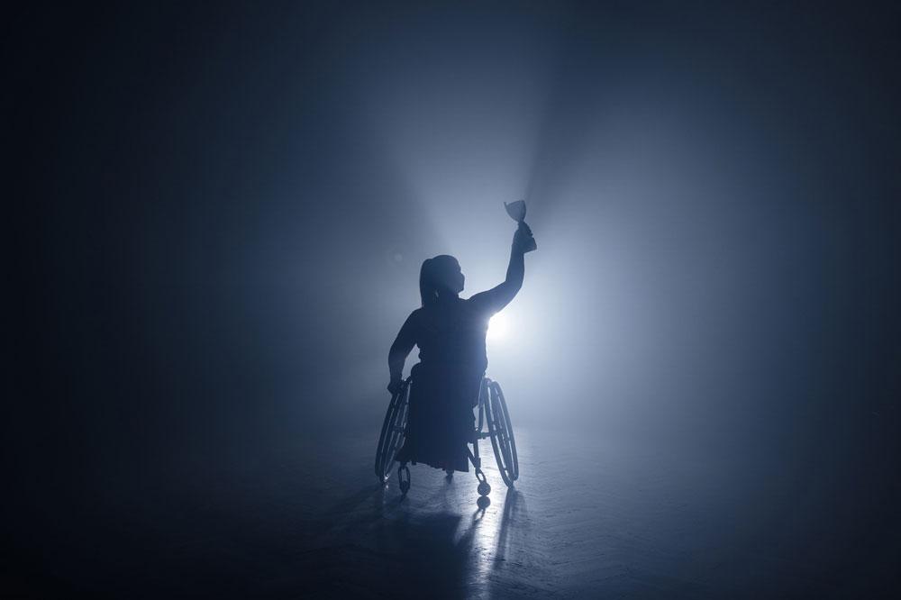 Female athlete in wheelchair in shadow holding up a trophy.