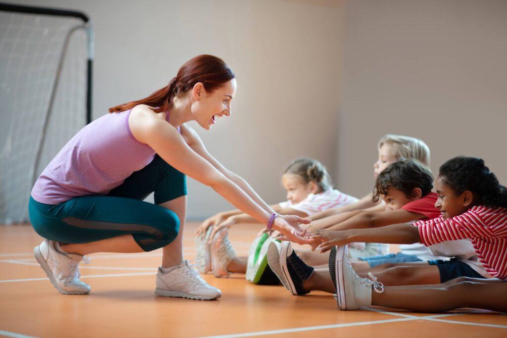 Young female teacher helping a diverse group of kids stretch in a gym.