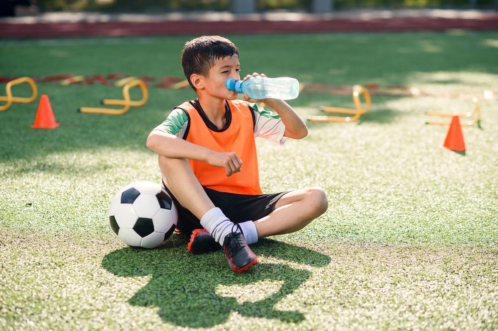 Young male soccer player sitting on the field drinking water.