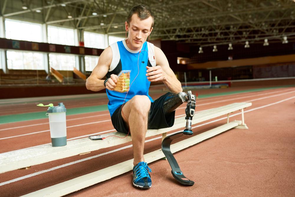 Male track athlete with a prosthetic leg eating a sandwich on a bench.