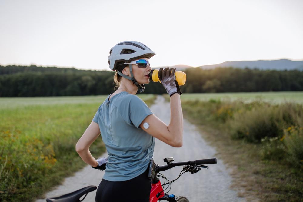 Young female cyclist drinking a water bottle wearing a glucose monitor on her arm.