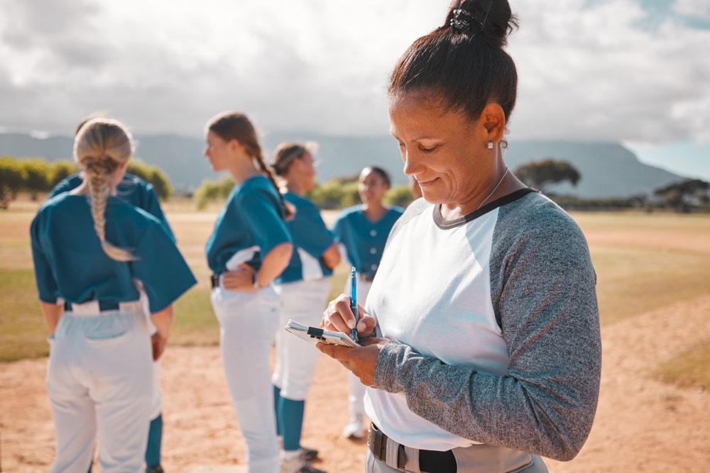 Female softball coach writing on pad in front of team.