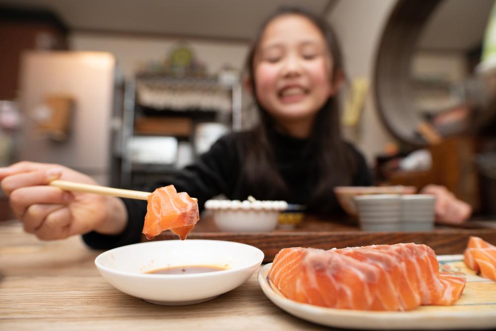 Young asian girl smiling while using chopsticks to pick up a piece of salmon.