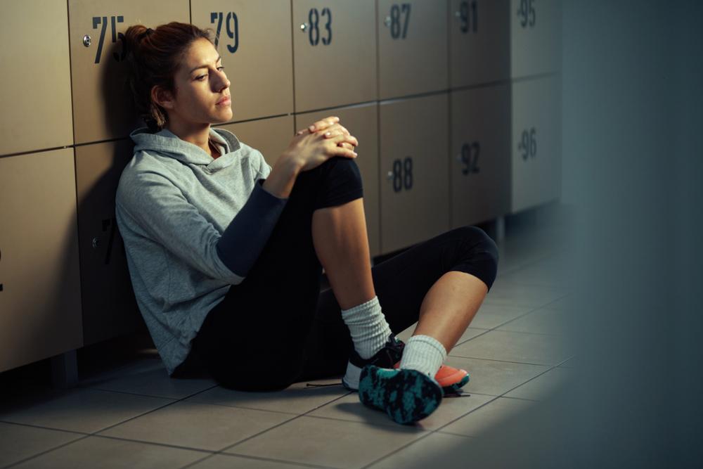 Teen girl sitting on floor in locker room looking dejected.