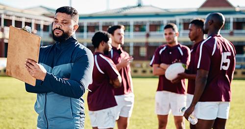 Rugby coach looking at clipboard next to team of teen boys.