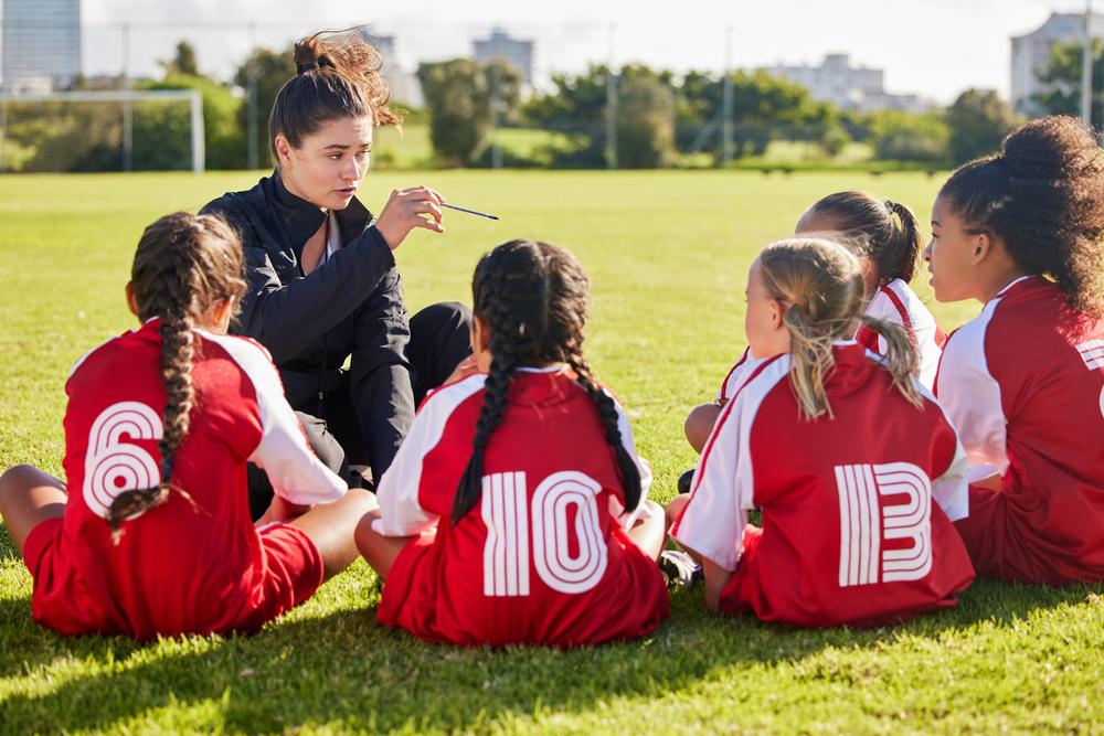 Female coach talking to young girl soccer team on field.