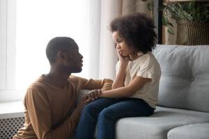 Black father talking to concerned daughter on couch.