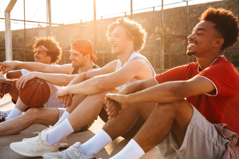 Diverse group of young men sitting on basketball court, laughing.