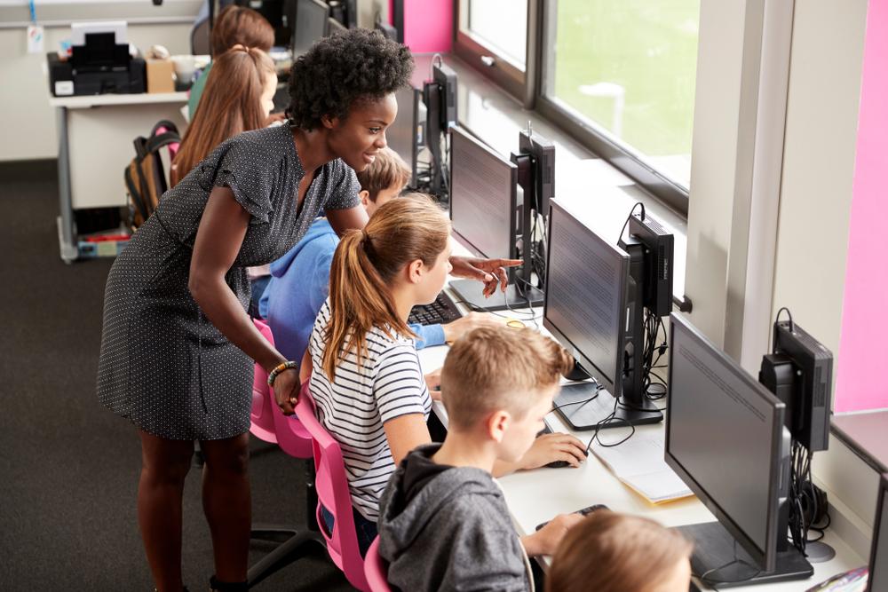Teacher in classroom assisting children with computer learning.