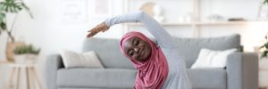 Young black woman wearing a hijab stretching in front of a laptop in her home.