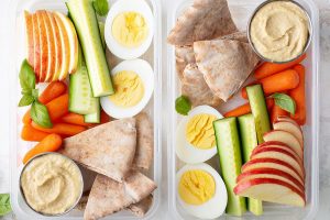 Two tupperware containers filled with veggies, hard boiled eggs, pita, hummus, and sliced apples.