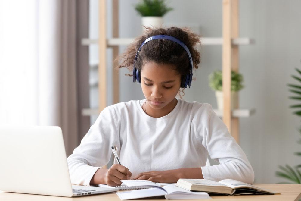 Teen girl writing in a note book while wearing headphones.