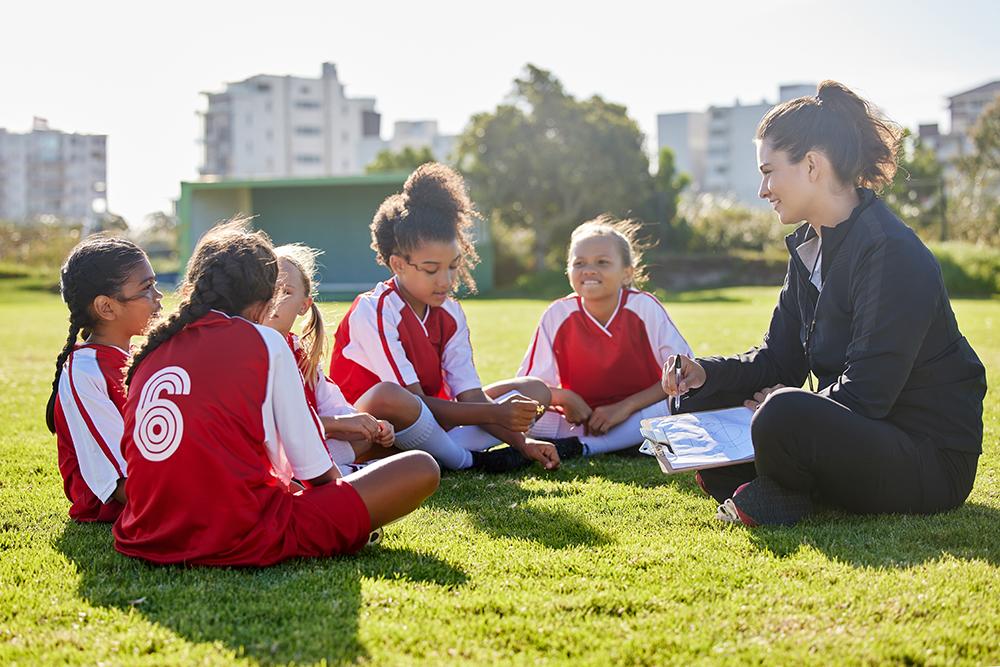 Female coach sitting on field with young girl soccer team.