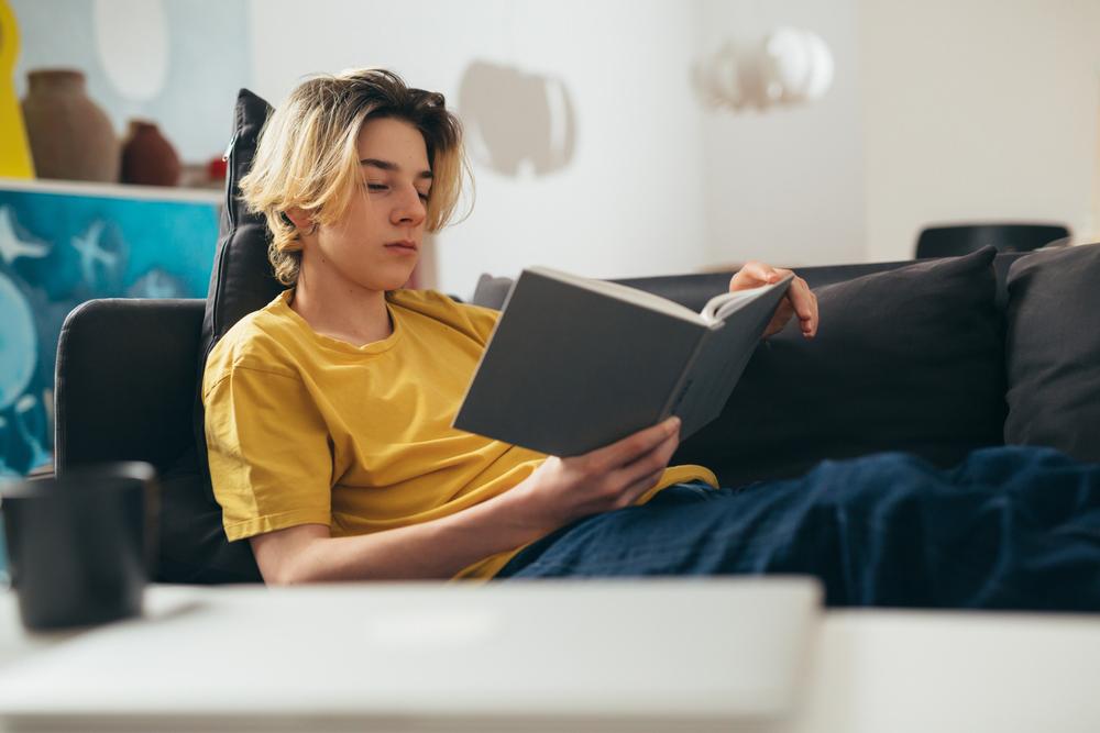 Teen boy reading a book on a couch.