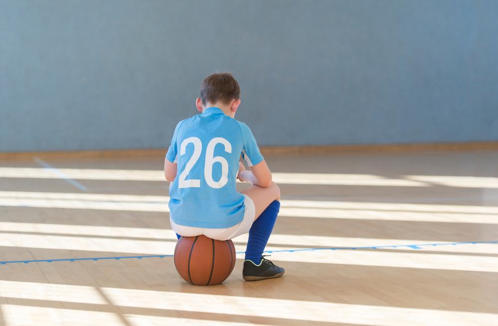A young male basketball player sitting on a ball turned away.