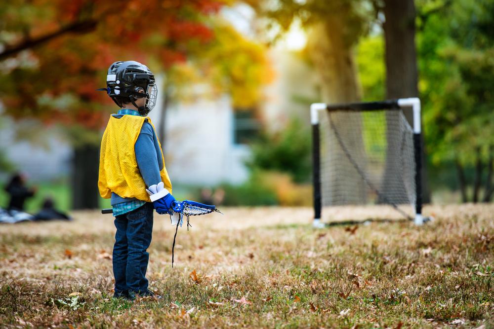 Young kid in lacrosse gear standing alone in front of a goal.