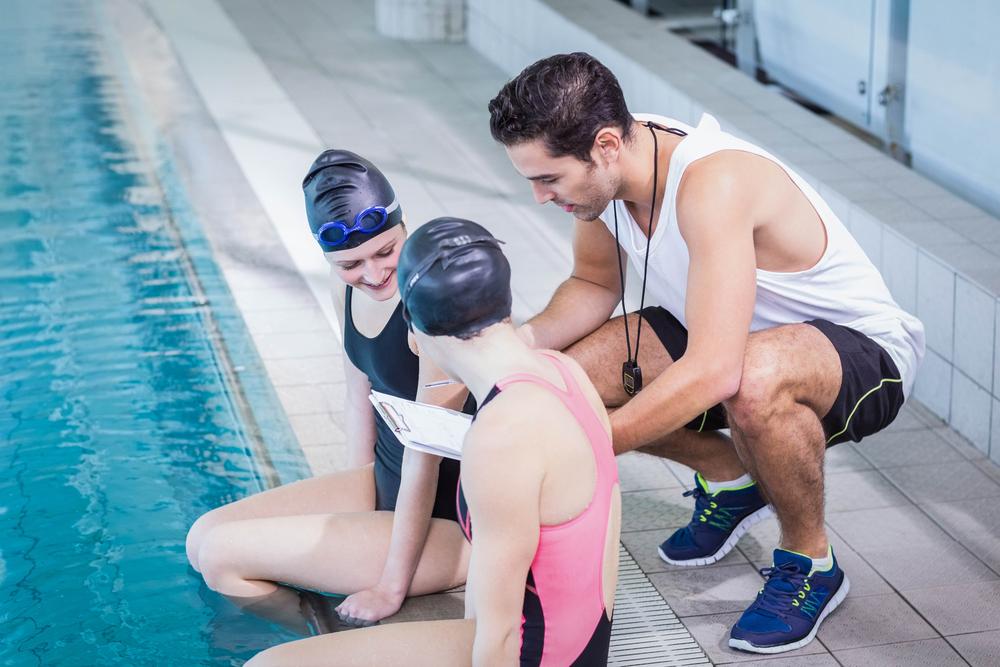 Male swimming coach talking to two young female swimmers next to pool.