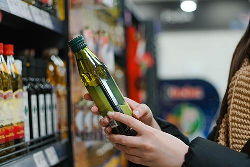 Hands holding a green olive oil bottle in a grocery store aisle.