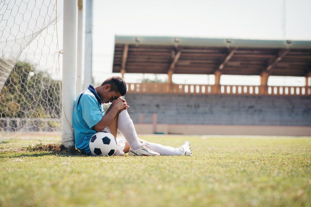 Young male soccer player sitting against goal post looking down.