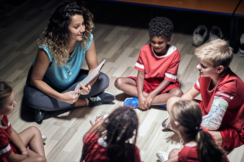 Adult woman talking to diverse soccer team indoors.