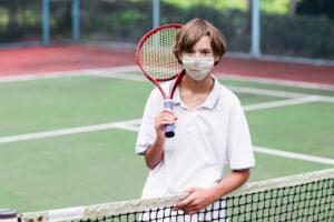 Young teen male wearing a fask mask holding a tennis racket on a tennis court.