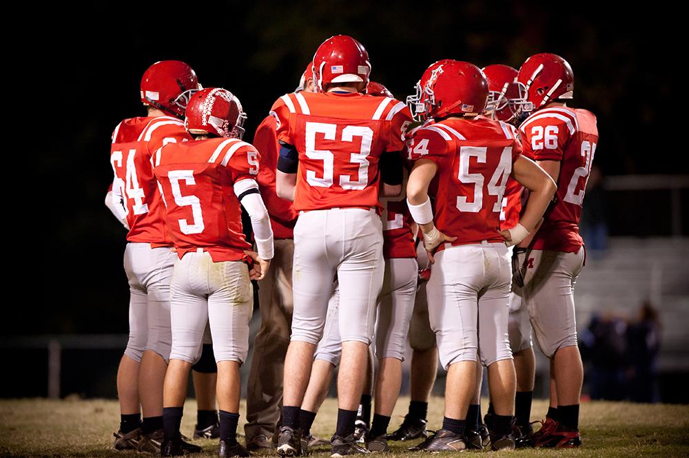 Football team huddled on field during night game.