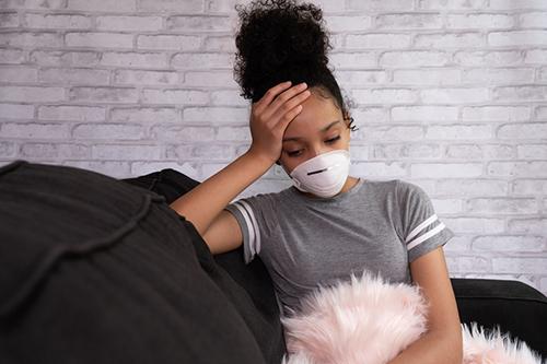 Young teen on couch looking down wearing a mask.