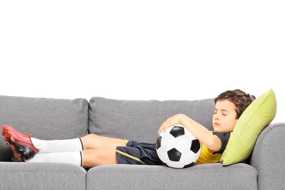 Boy in soccer outfit with ball sleeping on couch.