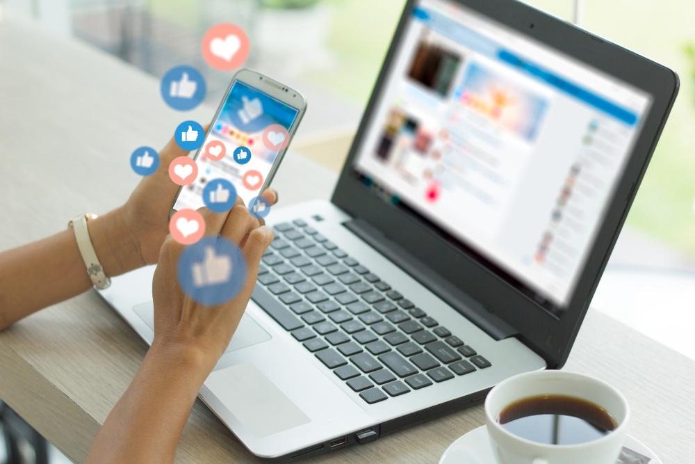 Young woman's hands holding a smart phone and looking at a laptop with social media icons.