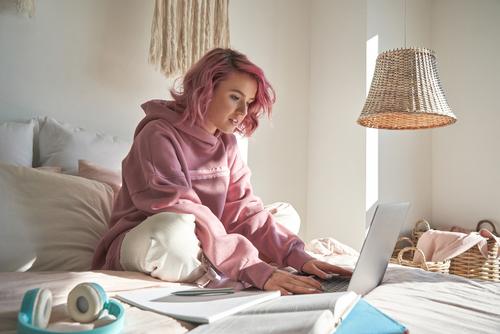 Young teen with pink hair on laptop in bed.