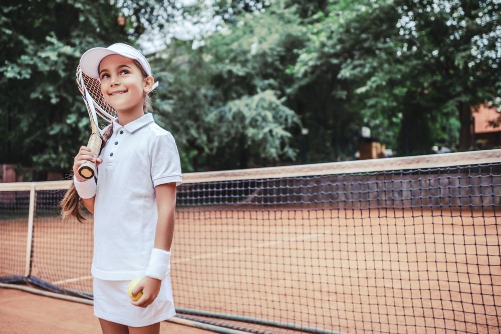 Little girl smiling on a tennis court in tennis gear, holding a racket.