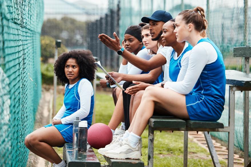 A male coach with his diverse young female sports team in the stands.