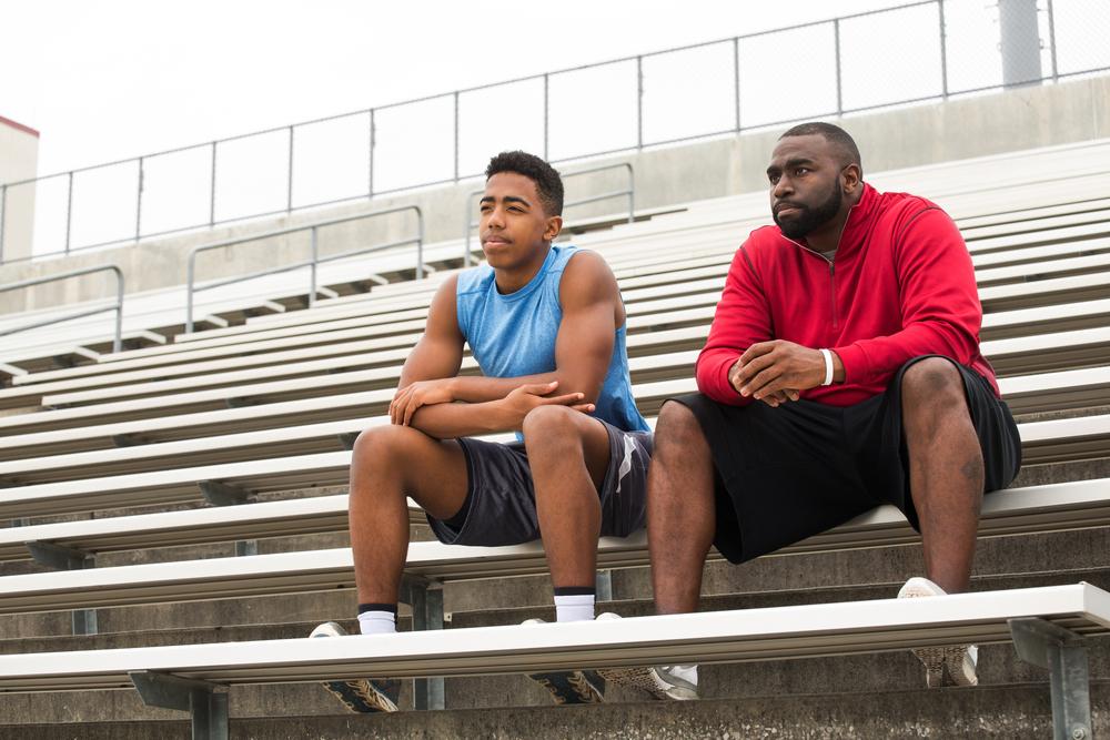 Coach sitting alone with teen male in football stadium stands.
