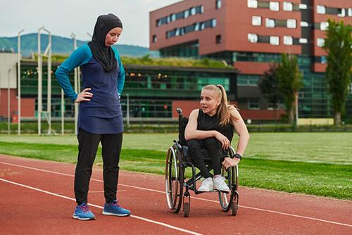 Woman in wheelchair on track talking to another woman wearing a hijab.