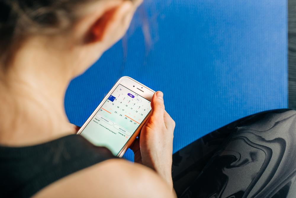 Woman looking at calendar tracking app on phone on yoga mat.