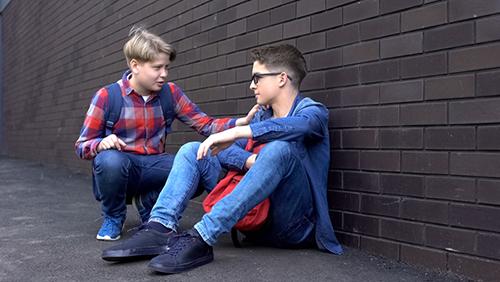 Two young white teens talking against a wall.